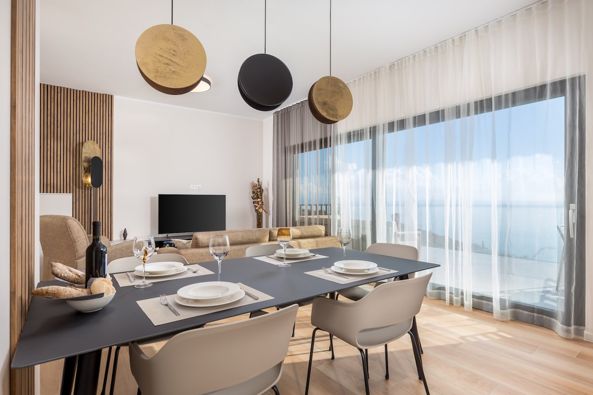 A dining area features a modern table set for four, complemented by stylish chairs. Above, circular light fixtures add warmth. A large window offers panoramic views of the sea, while sheer curtains softly diffuse natural light, enhancing the spacious feel of the room.
