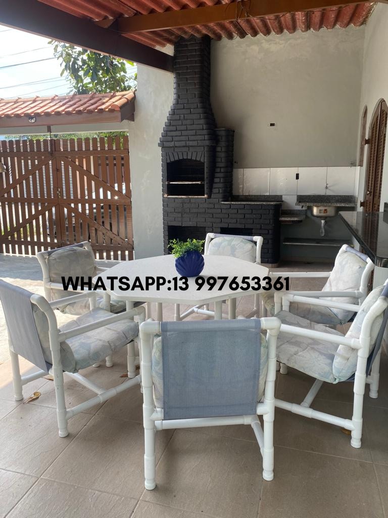 An outdoor dining area is featured with a round table surrounded by six chairs, complemented by a cozy barbecue grill. The setting showcases neutral tiles and stone walls, providing an inviting space for gatherings. Sunlight filters through the overhead structure, enhancing the relaxed atmosphere.