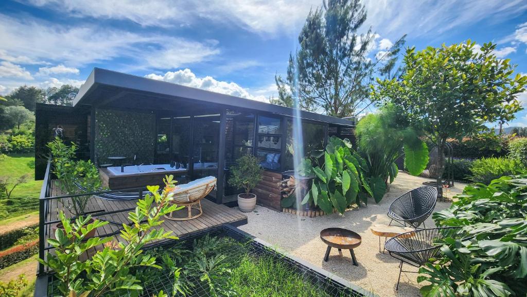 A modern cabin design is nestled in a lush landscape, featuring a spacious deck bordered by vibrant greenery. Comfortable seating arrangements, including round chairs and a fire pit, provide an inviting outdoor space for relaxation. The sky above is bright with scattered clouds.