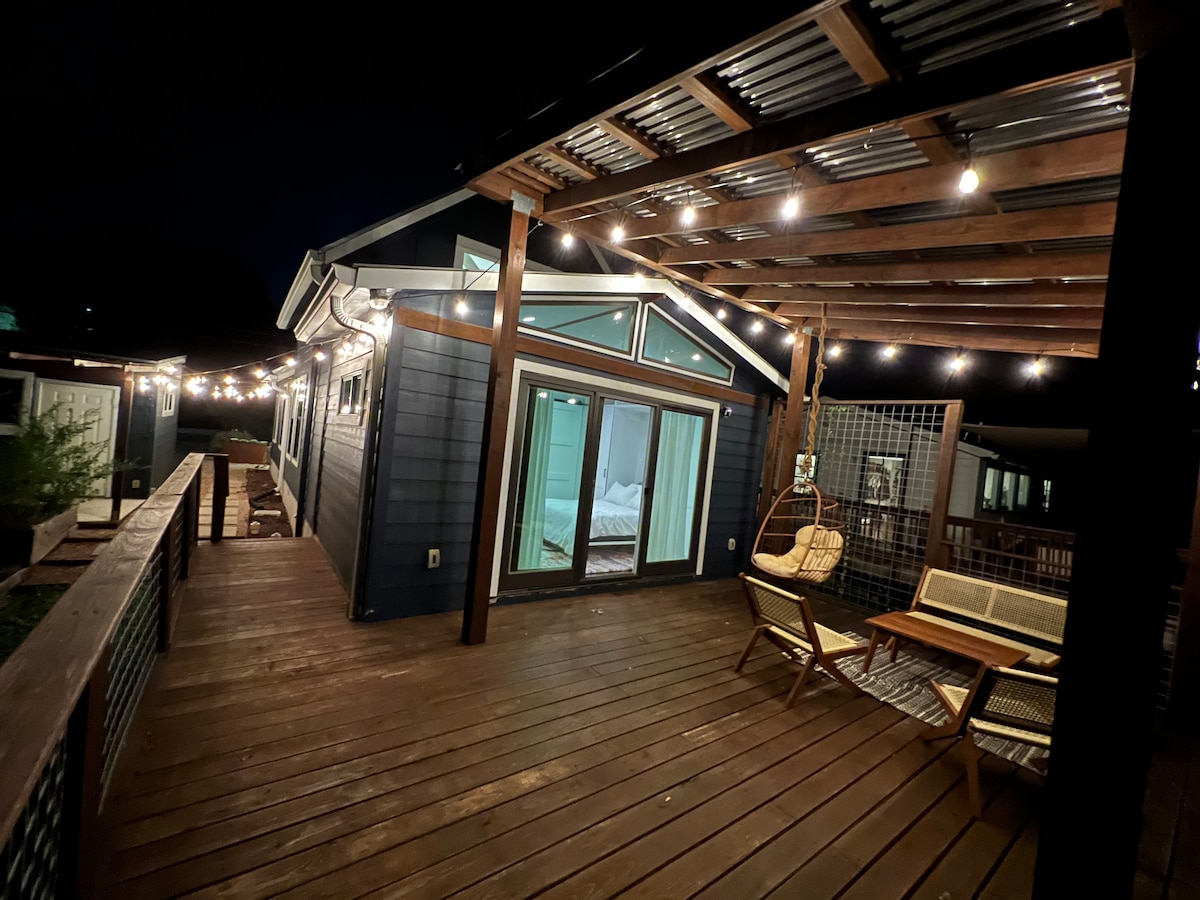 A wooden deck is illuminated by string lights, creating a warm ambiance. The modern house features large glass doors, leading to an interior space. Patio chairs are arranged invitingly, surrounded by a tranquil setting that blends with the evening sky.