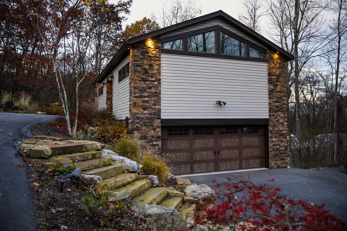A modern guest house is situated on a sloped driveway, featuring a blend of stone and siding on its exterior. Large windows allow natural light to fill the space. A landscaped pathway leads to the entrance, framed by trees and shrubs.