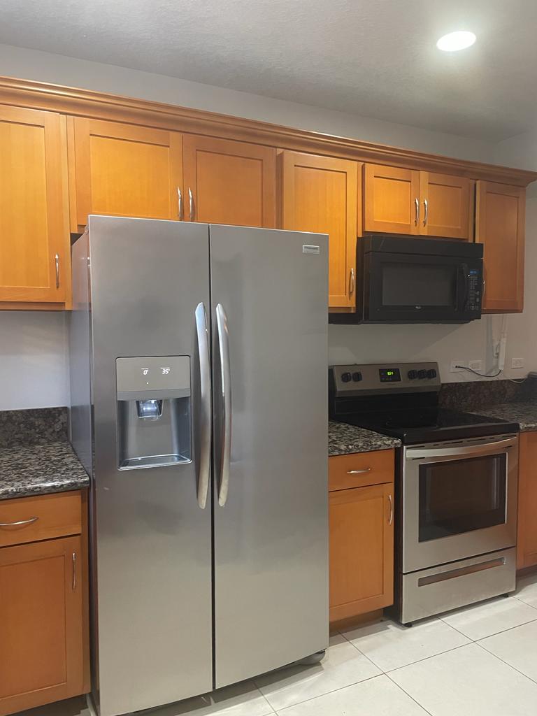A modern kitchen features wooden cabinetry and sleek granite countertops. A stainless steel refrigerator stands alongside a built-in microwave and an oven with a stovetop. Soft lighting enhances the kitchen's functional layout, providing a welcoming space for meal preparation.