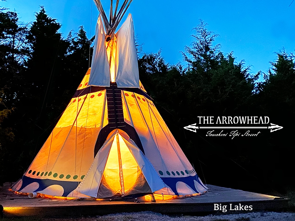 The tipi is illuminated from within, showcasing its unique design and vibrant colors. The structure stands prominently on a wooden platform, surrounded by natural greenery, creating a welcoming and distinctive glamping experience.