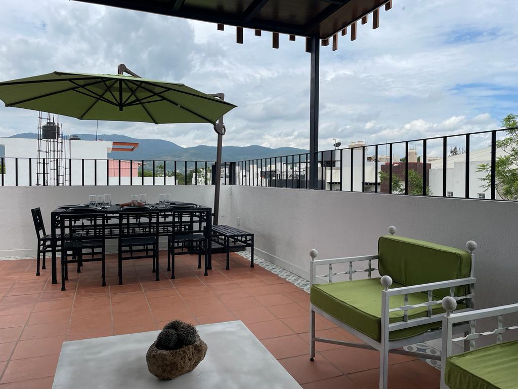 An outdoor terrace features a dining table with black metal chairs, shaded by a green umbrella. Green seating areas with cushions complement the space, surrounded by a scenic view of the mountains in the background. The tiled floor adds a distinctive texture to the setting.