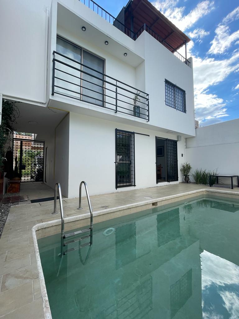 A private pool area is visible, featuring a clean water surface reflecting the sky. Adjacent to the pool, a modern two-level building is partially shown, with large windows and a balcony overlooking the space. Lush greenery adds a touch of nature to the environment.