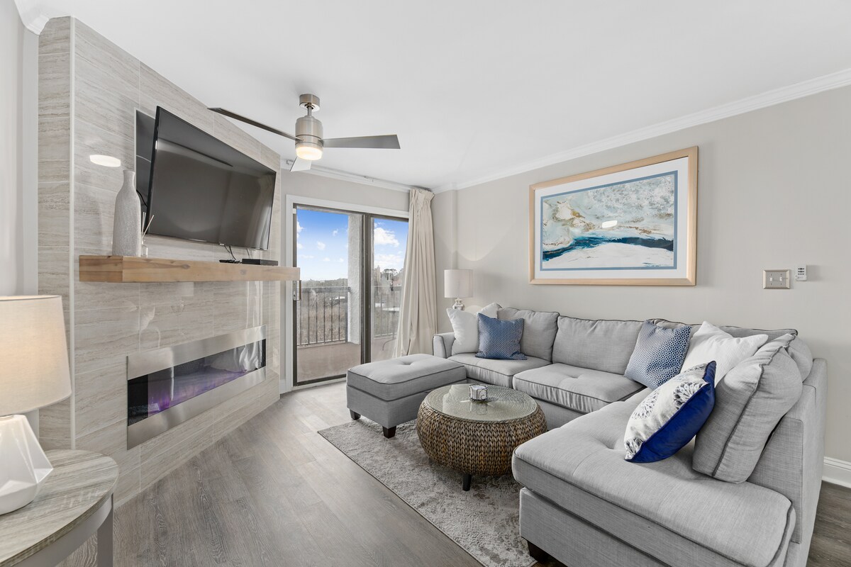The living room showcases a spacious sectional sofa in soft gray, complemented by a decorative pillow. A modern TV is mounted above a sleek fireplace, and large glass doors provide access to the balcony, allowing natural light to fill the space.