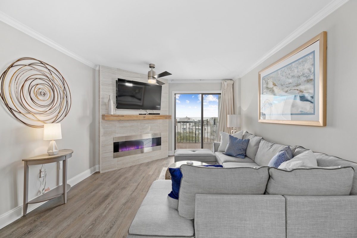 The living area features a modern design with a large gray sectional sofa, offering ample seating. A wall-mounted flat-screen TV is positioned above a sleek fireplace. Natural light fills the space through large sliding glass doors, leading to the balcony with a view.