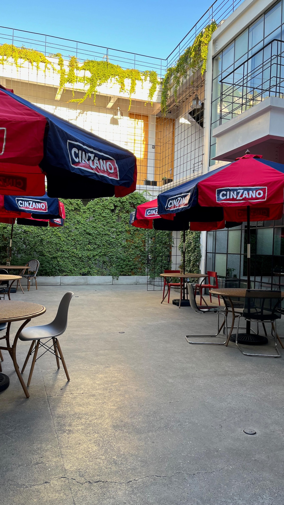 An outdoor seating area features several round tables beneath large red umbrellas. The space is surrounded by greenery, with a vertical garden visible on one wall. Natural light illuminates the concrete surface, creating a welcoming atmosphere.