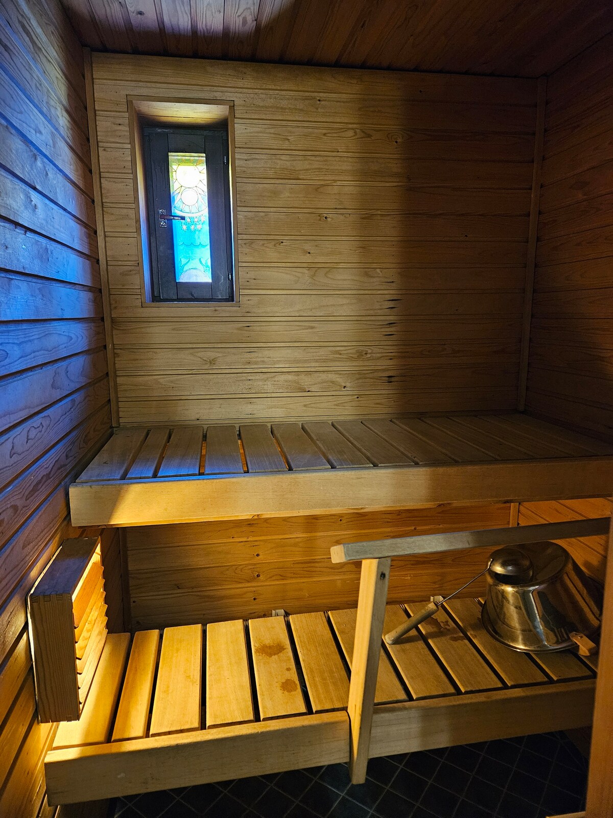 A wooden sauna features a bench structure along the walls, designed for relaxation. A small stained glass window allows natural light to enter, illuminating the warm, wooden interiors. A metal water vessel is positioned on one of the benches, ready for use.