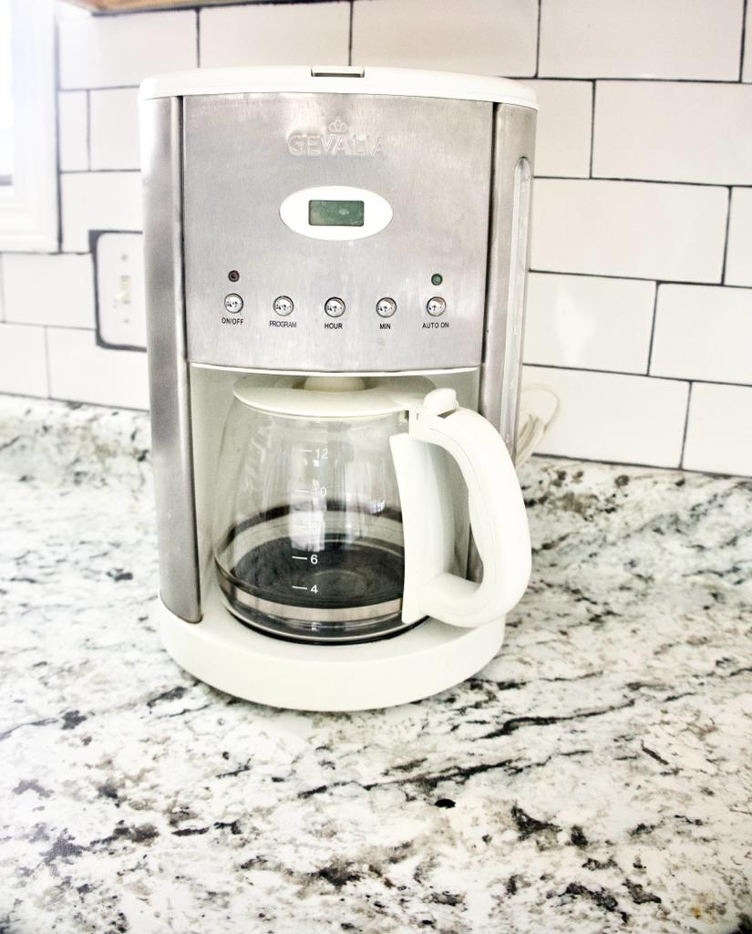 A coffee maker is positioned on a granite countertop, featuring a stainless steel exterior and a glass carafe. The control panel at the top displays buttons for various brewing options, and the coffee maker is situated near a bright kitchen window.