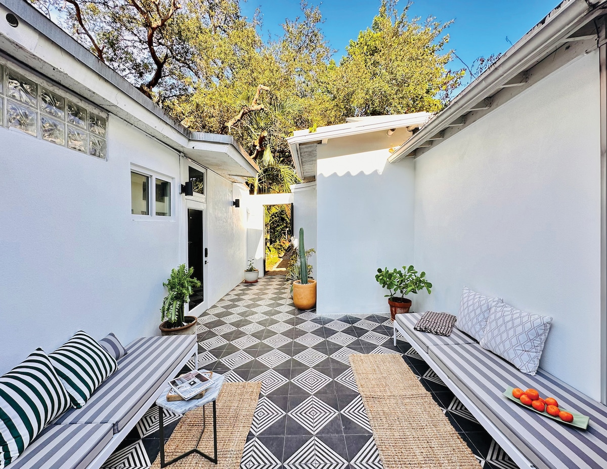 A private courtyard features patterned tiles and a bright, open layout. Two loungers are positioned on either side, adorned with striped cushions. Potted plants add greenery, while a small table holds fresh fruit, all surrounded by walls that invite natural light.