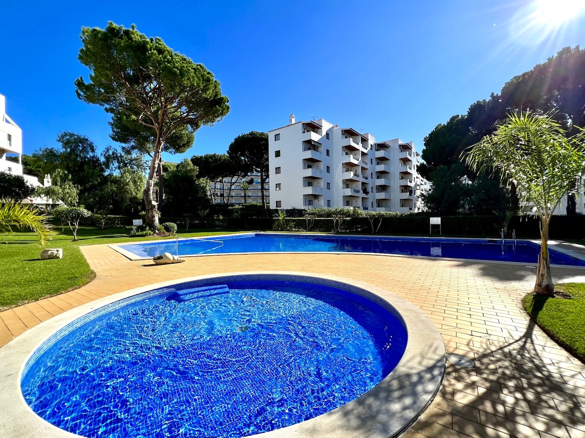 A serene pool area is presented, featuring a larger swimming pool and a smaller circular hot tub. Lush greenery surrounds the pools, and a residential building is visible in the background. The sky is clear and bright, reflecting in the water.
