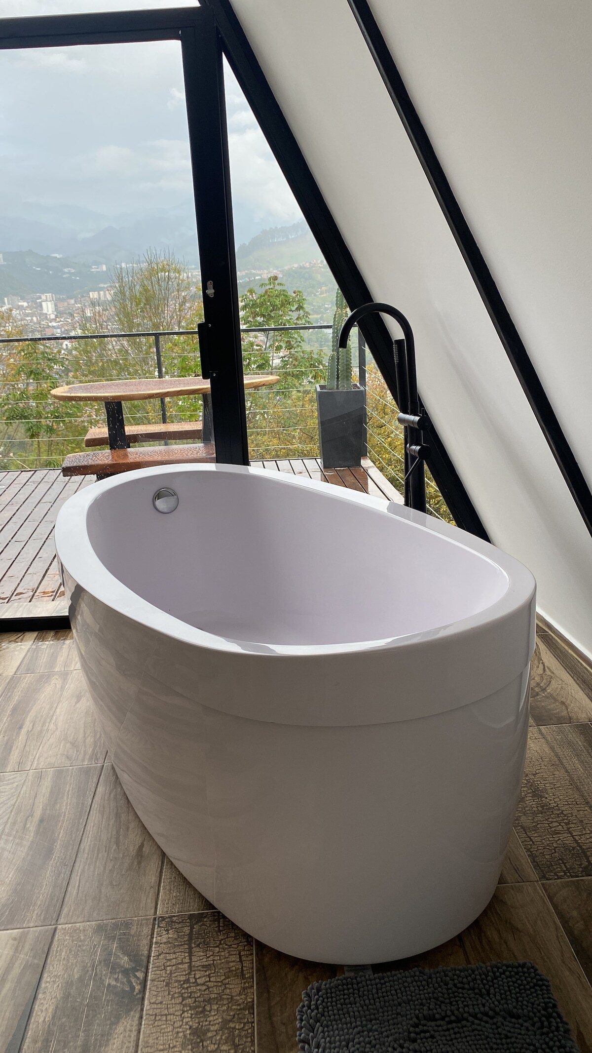 A modern, freestanding bathtub is positioned near a large window, offering expansive views of the surrounding nature and city. Natural light floods the space, highlighting the sleek design of the tub and the contemporary fixtures nearby. The floor is adorned with wooden tiles.
