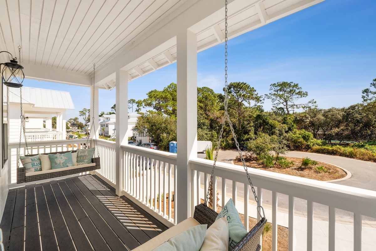 A covered porch features two swings with plush cushions, positioned under a white ceiling. Lush greenery is visible in the background, and a smooth road curves to the left. Bright, natural light illuminates the space, creating an inviting atmosphere for relaxation.