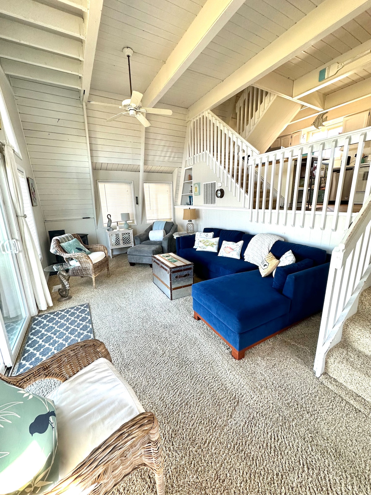 The living room features a spacious blue sectional sofa accompanied by a light gray armchair. A coffee table is positioned in the center, while natural light streams in through large windows. The vaulted ceiling enhances the airy atmosphere, and a ceiling fan is mounted overhead.