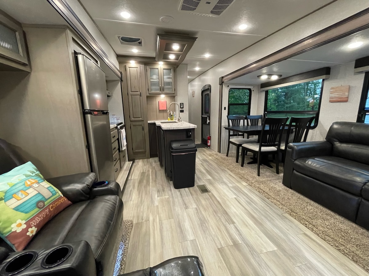 An open RV interior is displayed, featuring a well-equipped kitchen with modern appliances and a central island. A cozy living area is visible with two reclining chairs and a sofa, along with a dining space complete with a table and chairs. Natural light filters through large windows.