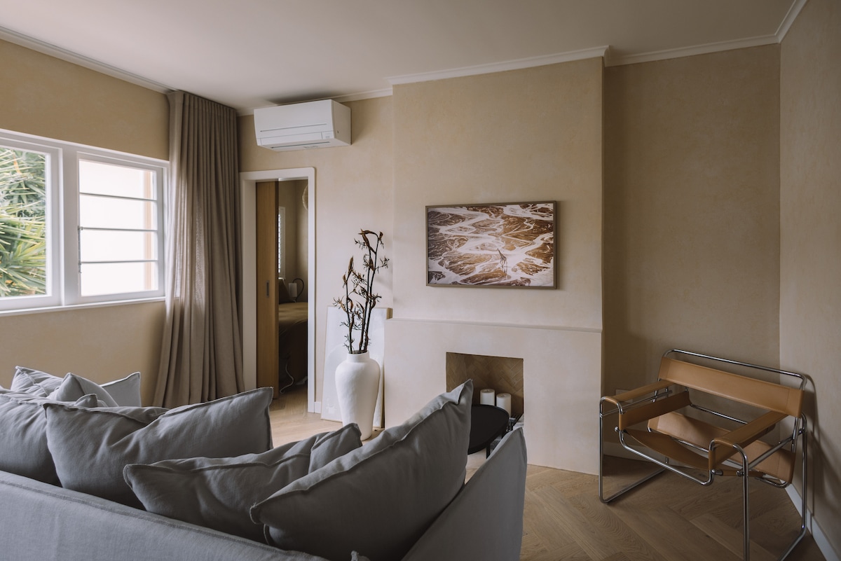 A cozy living area features soft grey sofas and a sleek leather chair. Natural light enters through a large window adorned with light curtains. An artwork depicting abstract waves hangs on the peachy limewashed wall, complemented by a minimalist vase with decorative branches.