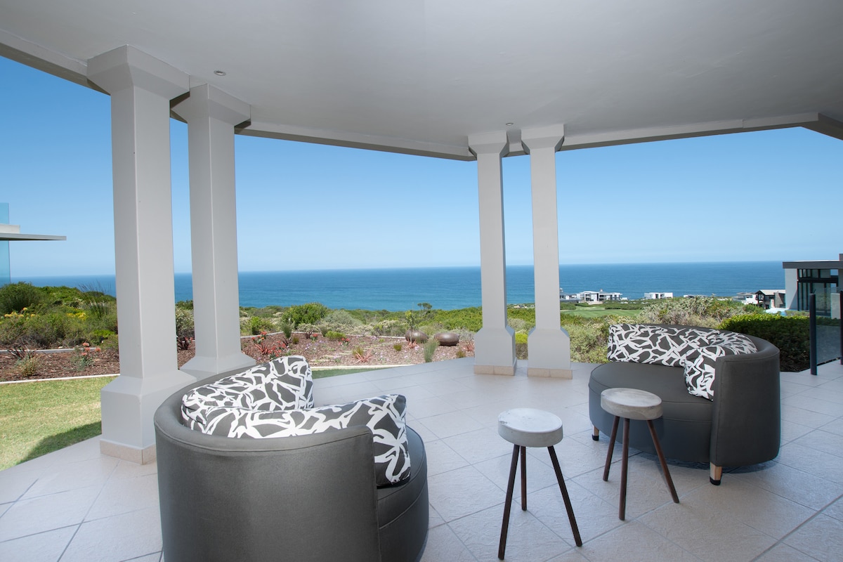 A covered terrace showcases two round seating areas with patterned cushions, positioned around small side tables. The scene is framed by columns, with sweeping views of the ocean and coastal landscape visible beyond the outdoor space.