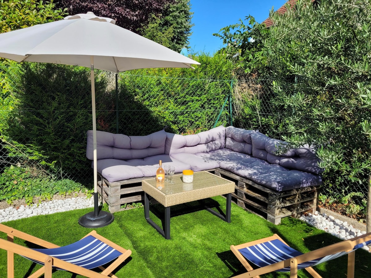 A private garden area features a cozy lounge set made from pallets, complemented by plush cushions. A table sits in the center, and a large umbrella provides shade. Surrounding greenery enhances the tranquil setting, with a small section of artificial grass visible underfoot.