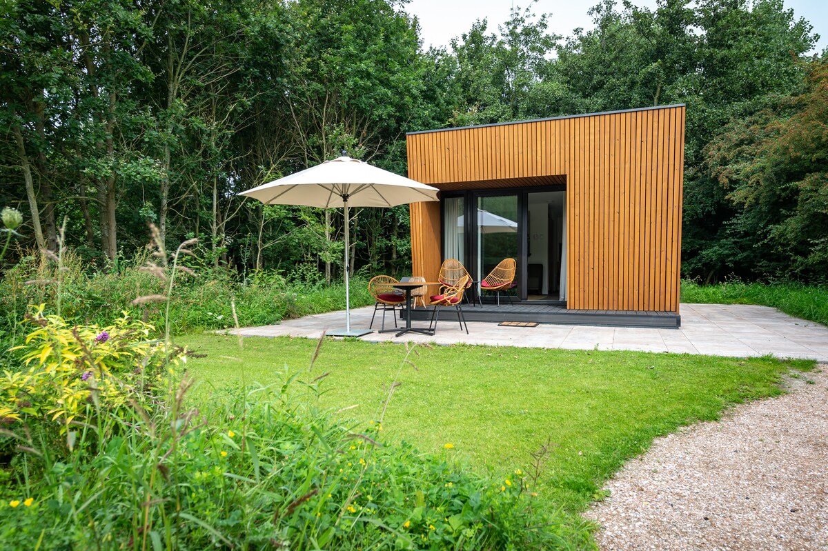 A modern lodge is situated in a lush natural setting, featuring wooden cladding and large glass doors. A patio with two wicker chairs and a small table is shaded by a large umbrella, surrounded by greenery and wildflowers.