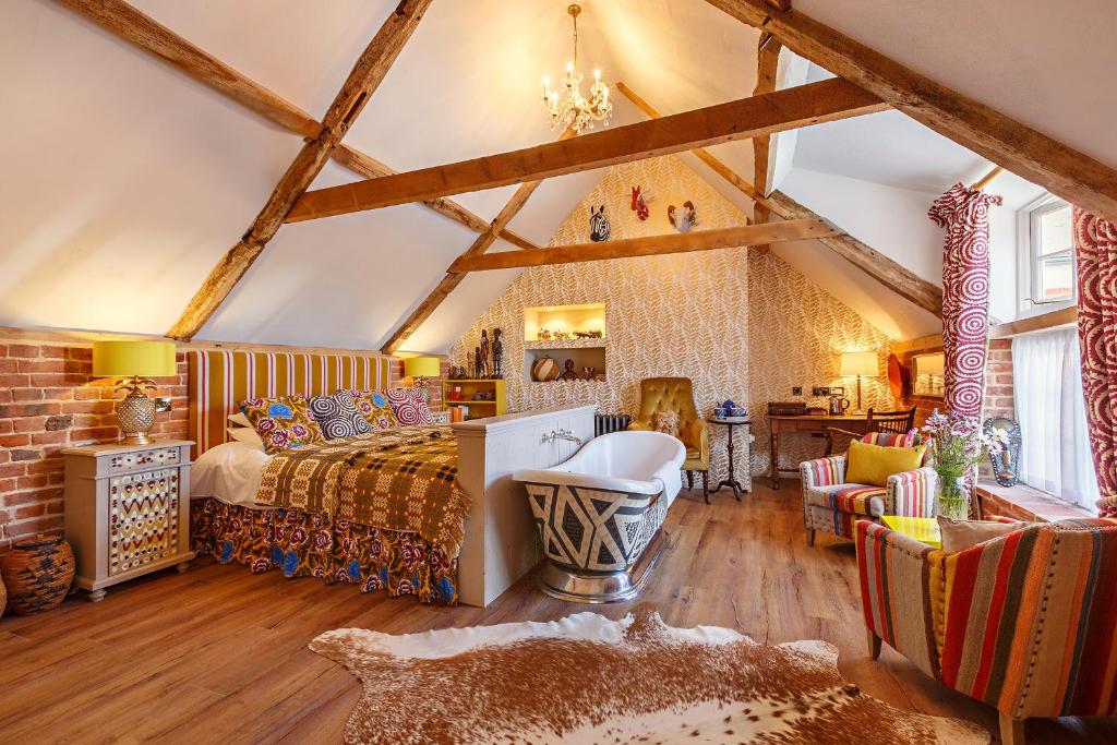 The spacious bedroom features wooden beams across the ceiling, complemented by a vintage chandelier. A roll-top bath and vibrant patterned fabrics are visible. Cozy seating is provided by two armchairs, and the decor includes animal motifs and colorful accents that echo African textiles.