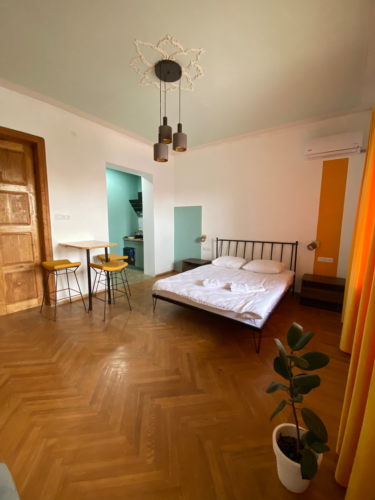 A well-lit attic studio features a spacious bed with fresh linens. Two bar stools sit at a small table beside a door leading to the kitchen area. A potted plant adds a touch of greenery, while warm yellow curtains frame a window, enhancing the airy feel.