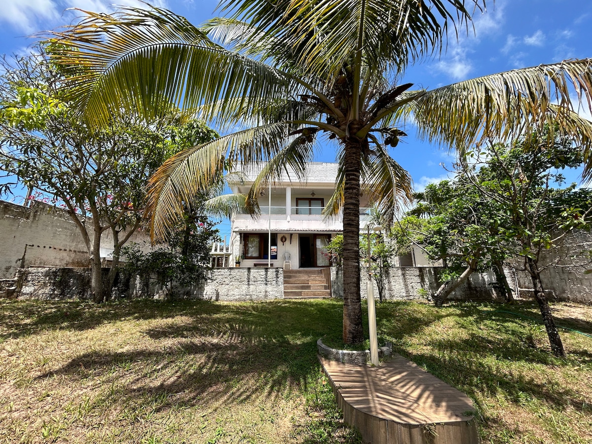 A two-story home is positioned among lush greenery, featuring a palm tree in the foreground. The structure is highlighted by a balcony and a staircase leading to the entrance. The landscaping is characterized by a grassy area and various trees, under a clear blue sky.