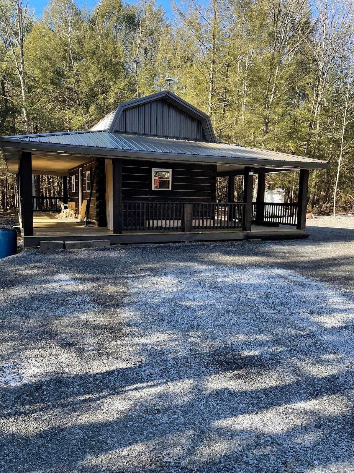 The log cabin features a charming front porch with a dark metal roof and wooden beams. Surrounded by trees, the structure is set on a gravel path. Sunlight creates patterns on the ground, highlighting the rustic design of the cabin.