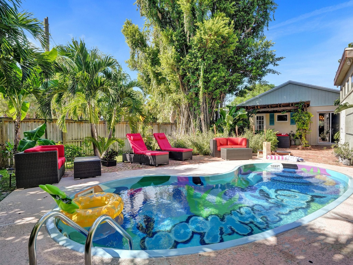 A vibrant backyard oasis features a kidney-shaped pool surrounded by tropical palm trees. Lounge chairs in bright red provide comfortable seating. Nearby, a covered seating area and a pool house create inviting spots for relaxation, complemented by lush greenery throughout the outdoor space.