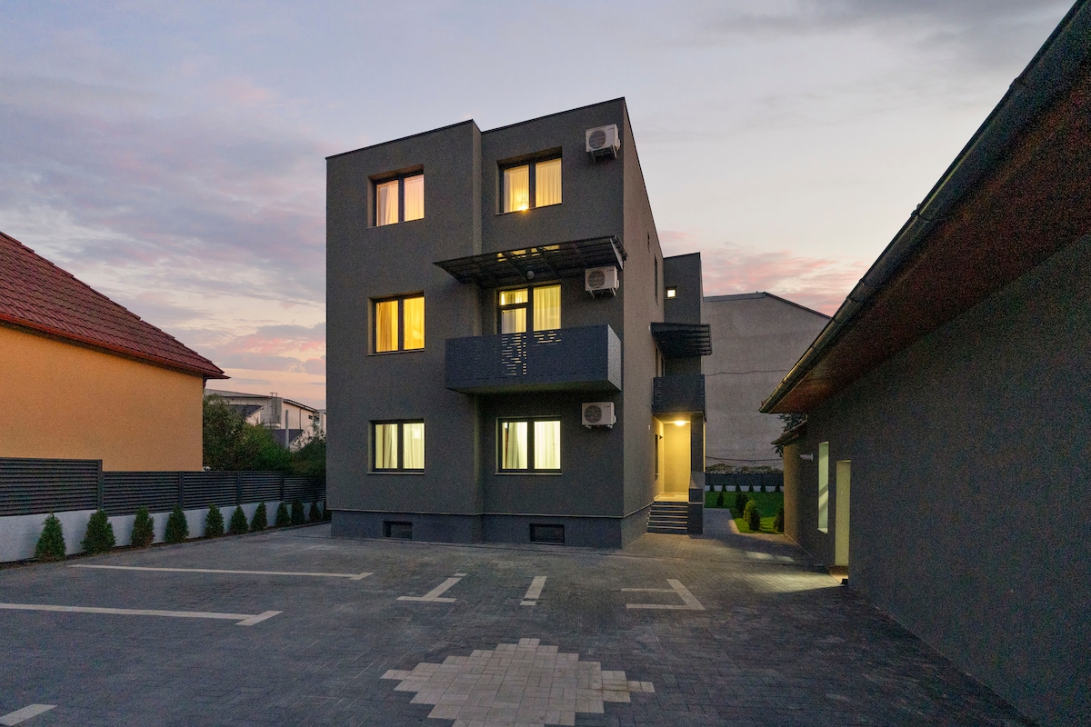 An architectural structure features a modern, gray exterior with multiple windows and a balcony. The building is situated in a spacious courtyard, surrounded by landscaping and adjacent to another single-story building. The scene is softly lit by the evening sky.