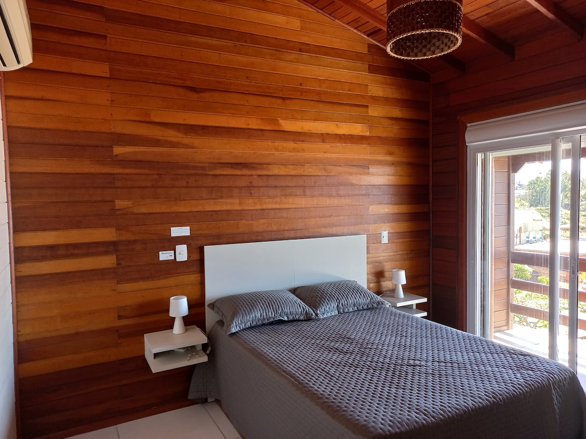 A tranquil bedroom features a wooden accent wall, creating a warm ambiance. A spacious bed is paired with two bedside tables, each equipped with a lamp. Large sliding glass doors provide access to outdoor views, allowing natural light to fill the room.