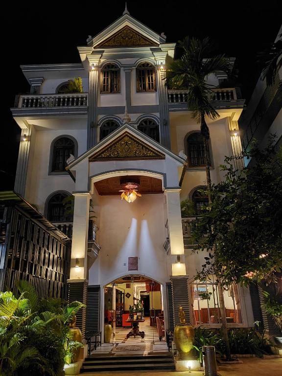 The exterior of PARENT HERITAGE ANGKOR VILLA is showcased at night, illuminated by warm lights. Architectural details include a triangular pediment above the entrance and decorative elements framing the large doors. Lush greenery is visible around the entrance, enhancing the inviting atmosphere.