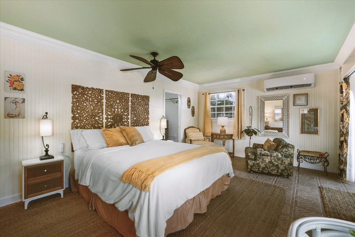 A spacious room features a large bed with a light-coloured coverlet, complemented by decorative wall art. A ceiling fan circulates air in the room. Natural light filters through windows, illuminating an armchair and a small sitting area. A calming green ceiling adds to the ambiance.