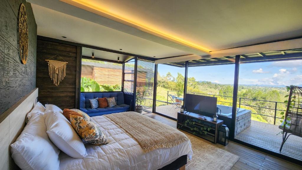 A spacious bedroom is displayed, featuring a large bed with neutral bedding and decorative pillows. A seating area with a sofa is present, and large glass doors lead to an outdoor deck with a jacuzzi, offering views of the surrounding landscape.
