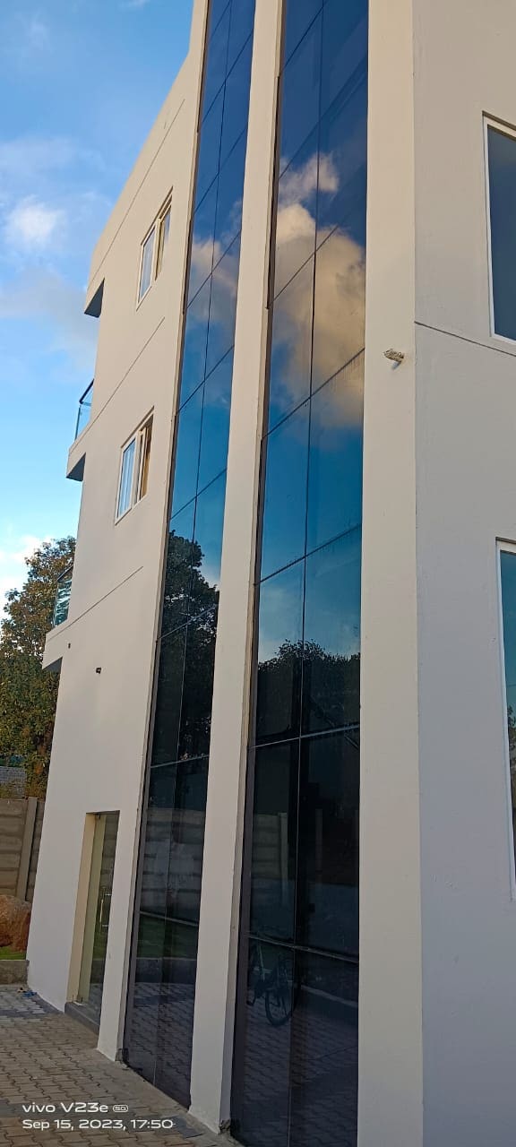 A modern building facade features sleek glass panels reflecting the surrounding trees and sky. The structure is predominantly white with minimalistic design elements, including several rectangular windows that enhance its contemporary appearance.