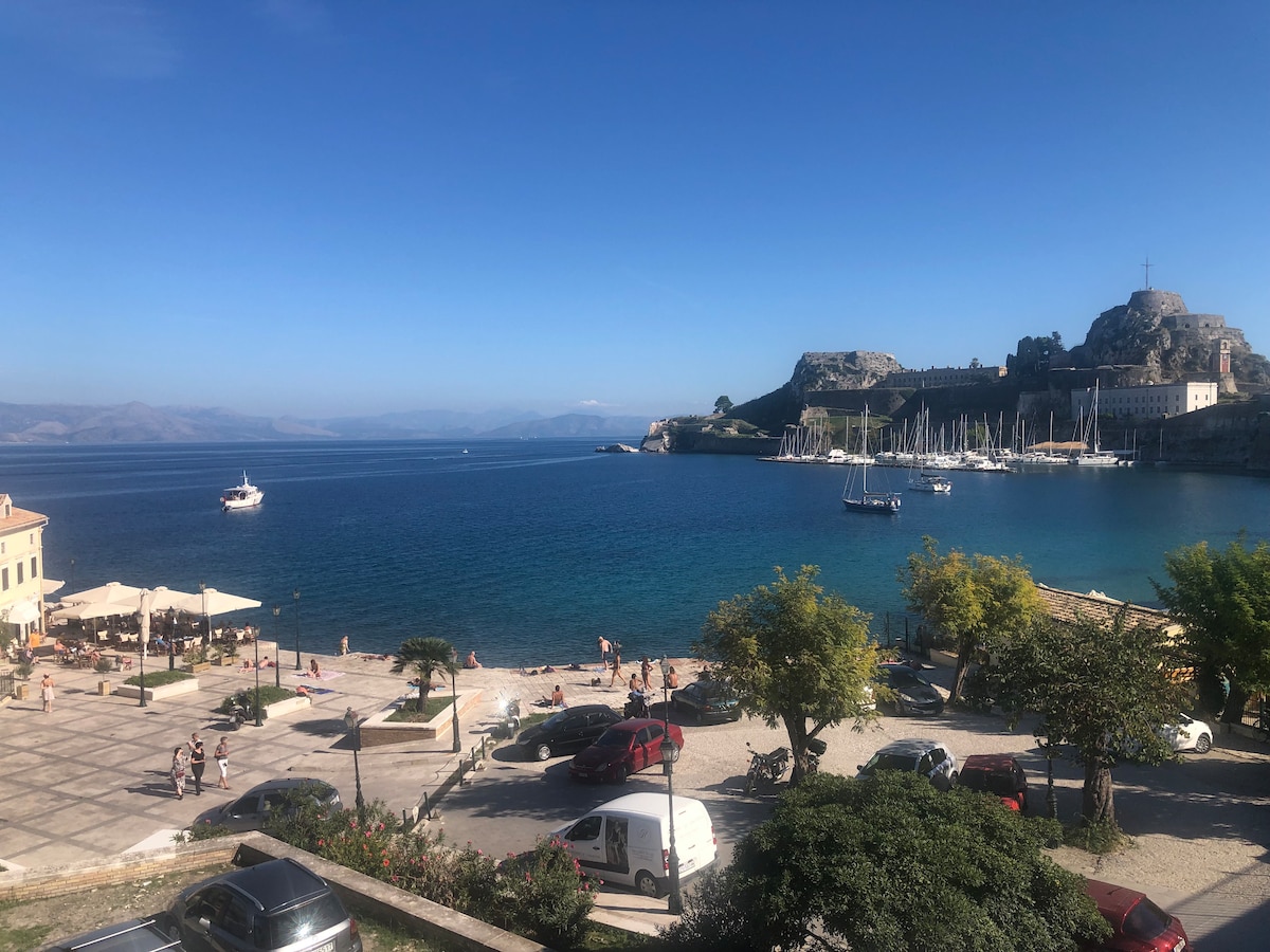 A serene view of the sea features calm blue waters and distant mountains. Boats are anchored in the harbor, with a bustling beach area visible. The scene includes a plaza lined with trees and outdoor seating under umbrellas, creating an inviting atmosphere for relaxation.