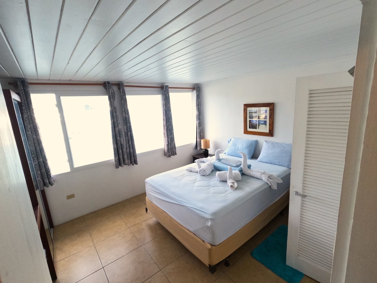 A well-lit bedroom features a spacious bed made with light blue linens and matching towels. Two windows provide natural light, and stylish curtains frame the views. A bedside lamp adds a touch of warmth, while light-colored walls create an airy atmosphere.