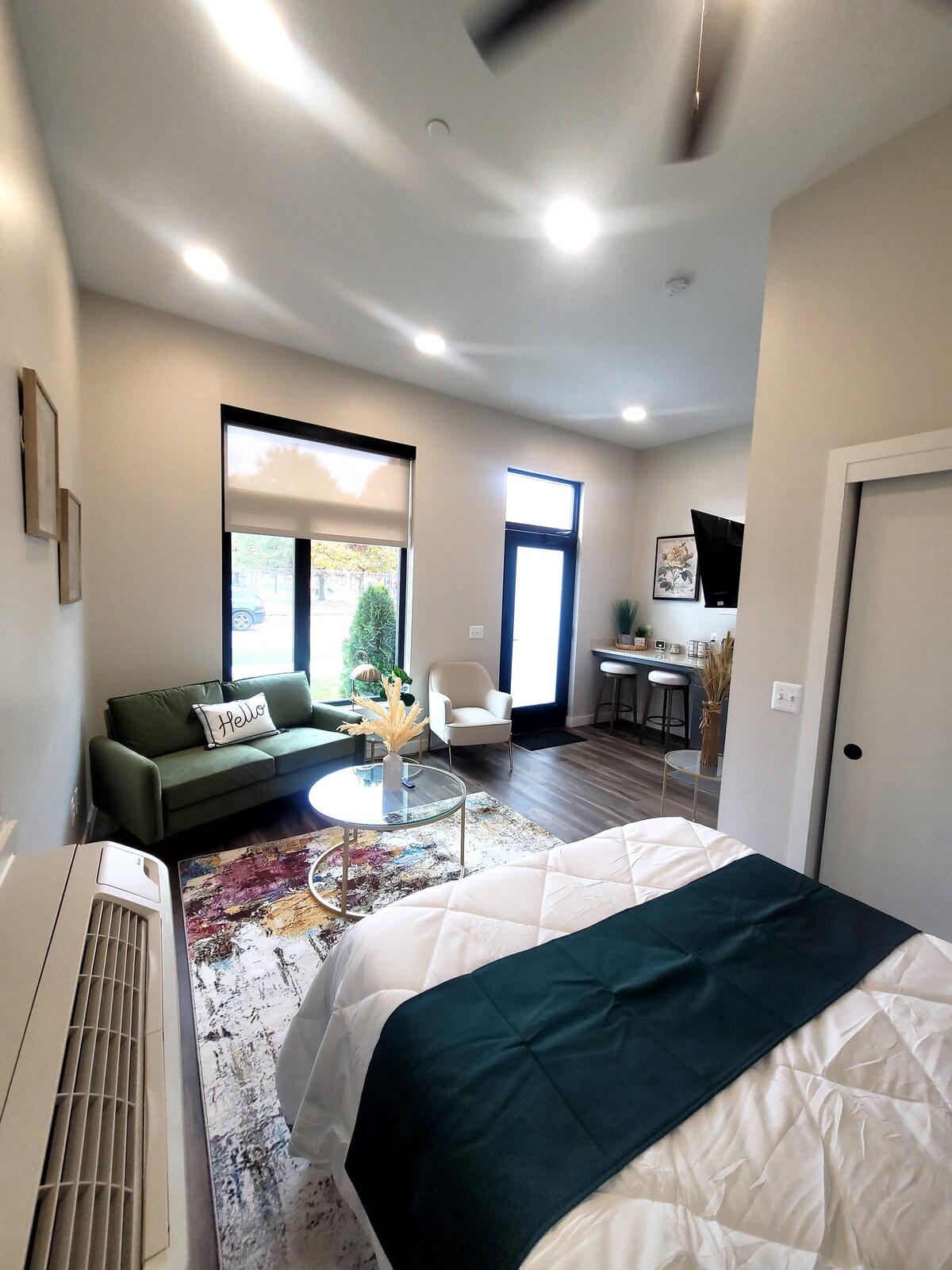 A well-lit room features a spacious bed with a textured blanket. A small green loveseat and a chair are arranged around a round table. Large windows allow natural light, while contemporary decor adds subtle style. A mounted TV is visible on the wall.