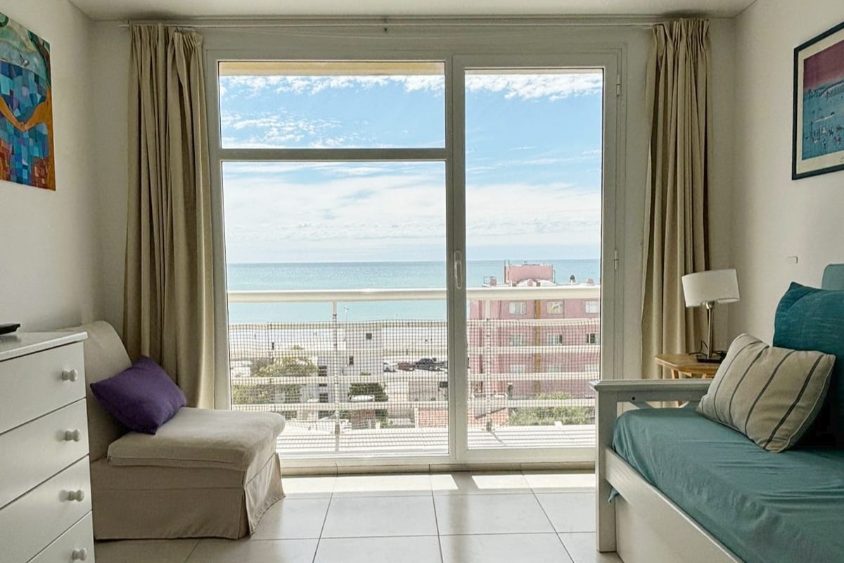 A bright living space features large glass doors that open to a stunning ocean view. Light-colored curtains frame the windows, and a cozy chair sits beside a white dresser. A turquoise bed with decorative pillows adds a pop of color to the room.