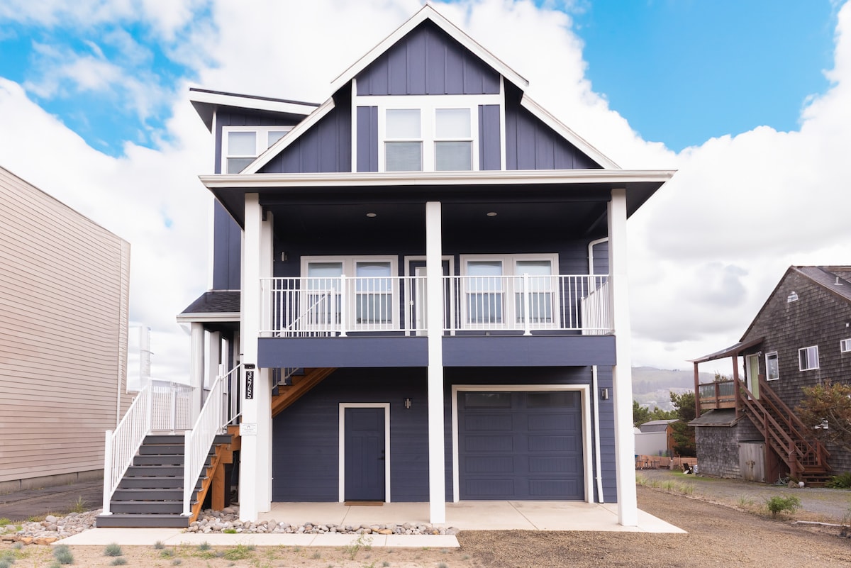 The exterior of a modern two-story home features a navy blue facade with white trim. A spacious porch is supported by pillars and overlooks a concrete driveway. Large windows provide ample natural light, while the surrounding area includes gravel and adjacent structures.