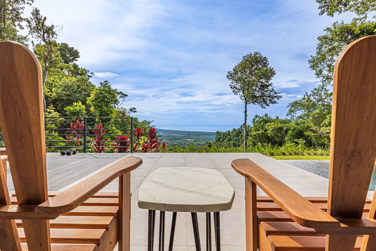 Two wooden chairs are positioned facing a scenic view, accompanied by a small, round marble-topped table. Lush greenery and vibrant plants surround the area, and a vast expanse of sky can be seen above, creating a serene outdoor setting.