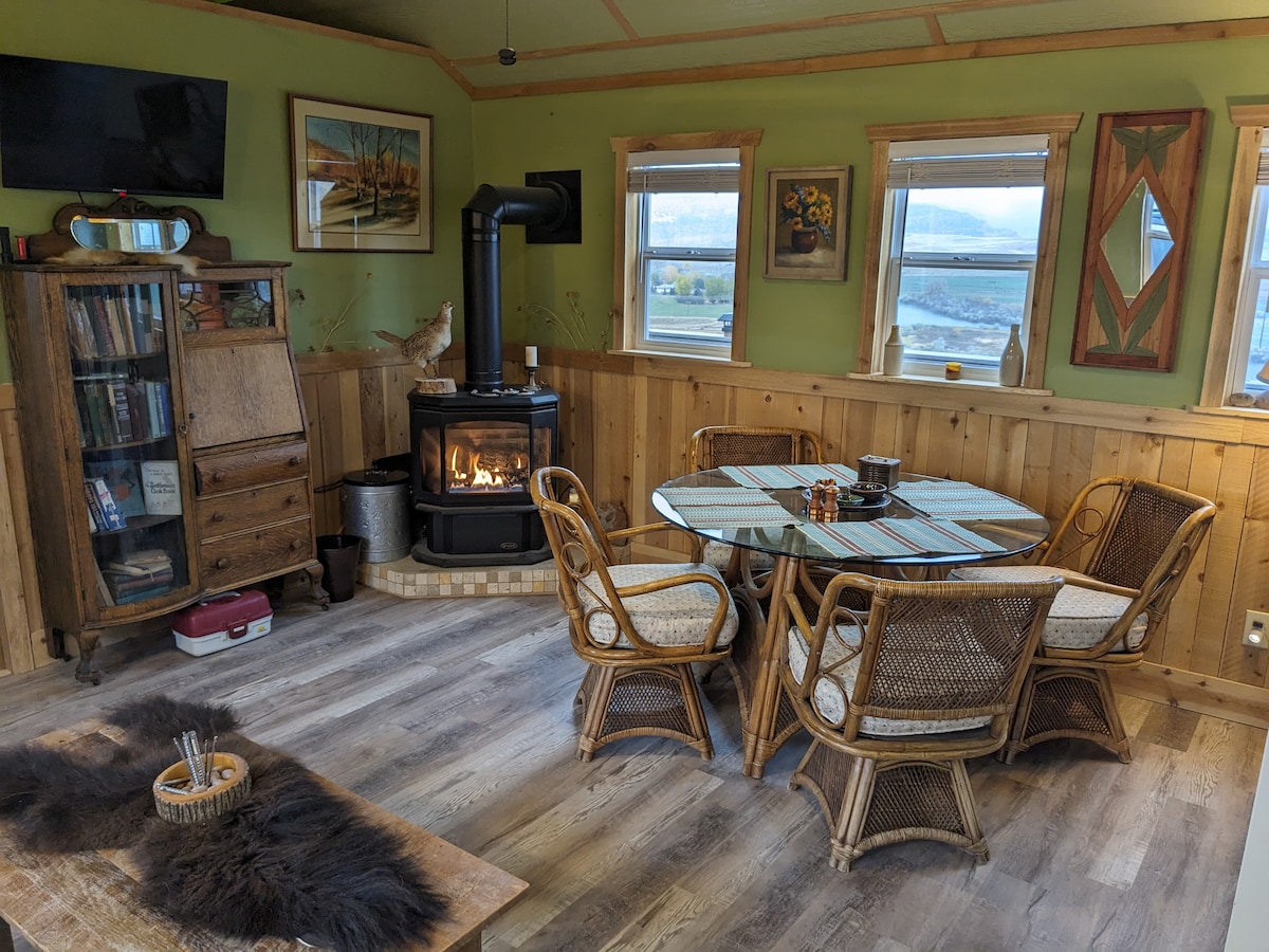 A cozy interior space features a round dining table surrounded by four woven chairs. A wood-burning stove adds warmth, while natural light floods in through the windows, revealing views of the surrounding landscape. Decorative art pieces adorn the light green walls.