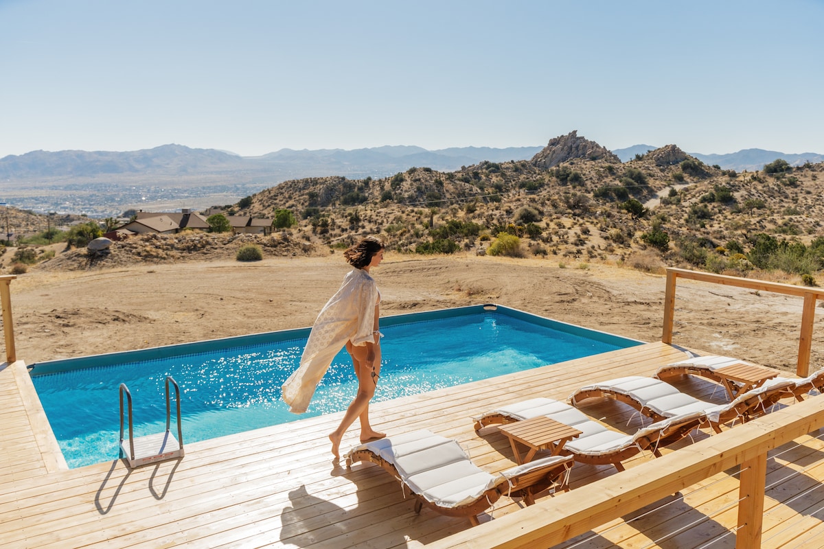 An inviting wooden deck surrounds a sparkling pool, with lounge chairs positioned for relaxation. Gentle hills and rocky outcrops create a scenic backdrop, emphasizing the open desert landscape under clear blue skies. A figure walks gracefully near the pool's edge, adding to the serene atmosphere.