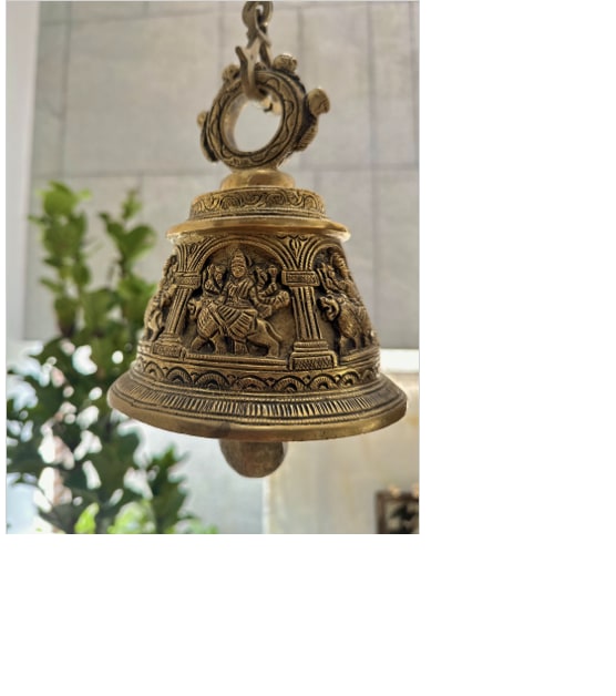 A detailed brass bell is displayed, featuring intricate carvings of deities around its base. The bell is suspended from an ornate loop. Green plants are softly blurred in the background, creating a contrast against the metallic sheen.
