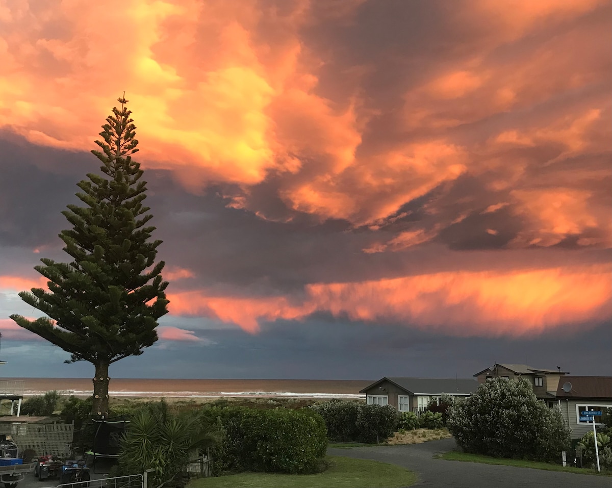 A striking sunset view is displayed, featuring vibrant orange and purple hues casting a warm glow across the sky. A tall evergreen tree stands prominently in the foreground, while the ocean is visible beyond the tree, reflecting the colorful sky.