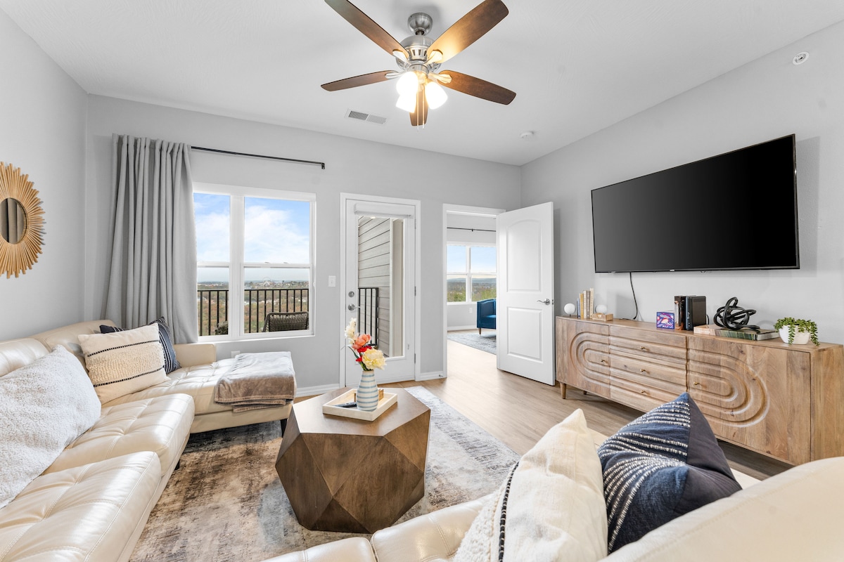 The living room features a leather sectional couch and a wooden coffee table with a decorative vase. A large flat-screen TV is mounted on the wall, and windows provide natural light and views, while a fan overhead adds comfort.