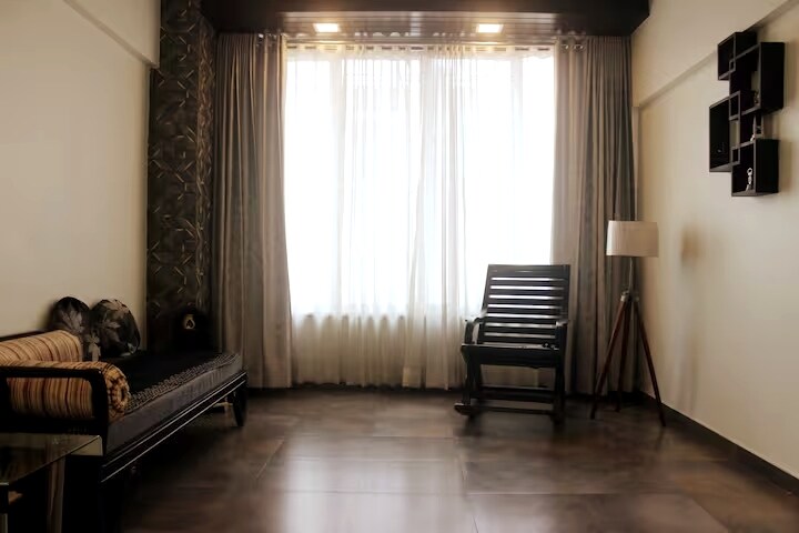 A bright living area is featured, characterized by large windows adorned with light gray curtains. A dark wood rocking chair is positioned next to a modern floor lamp. The wall showcases a minimalist shelf arrangement, while a textured accent wall adds depth.