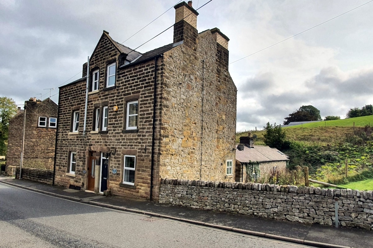 The stone-built cottage features a classic three-story design, with prominent chimneys and multiple windows that suggest an inviting interior. A low stone wall borders the property, with greenery visible in the background. The entrance is located at ground level, accessible from the street.