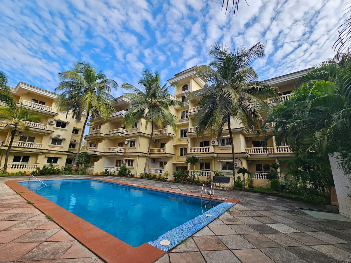 The spacious outdoor area features a large swimming pool surrounded by manicured gardens. The four-story building showcases a blend of balconies and windows framed by palm trees, with a partly cloudy sky above, creating a serene atmosphere.