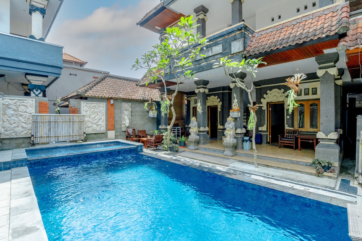 The outdoor area features a refreshing pool surrounded by stone pathways and tropical greenery. The architecture reflects traditional Balinese design, with decorative pillars and carvings. Comfortable seating is positioned nearby, inviting relaxation in a serene environment.